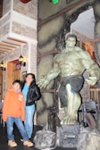 A large statue of a muscular, green-skinned superhero stands in an indoor setting. Next to the statue are a woman wearing a leather jacket and a young boy in an orange hoodie. The room is decorated with stone-like walls and vintage-style lamps.
