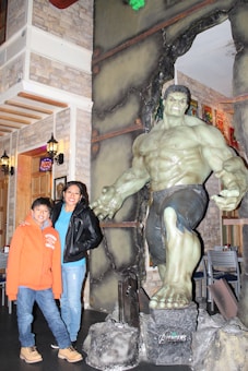 A large statue of a muscular, green-skinned superhero stands in an indoor setting. Next to the statue are a woman wearing a leather jacket and a young boy in an orange hoodie. The room is decorated with stone-like walls and vintage-style lamps.