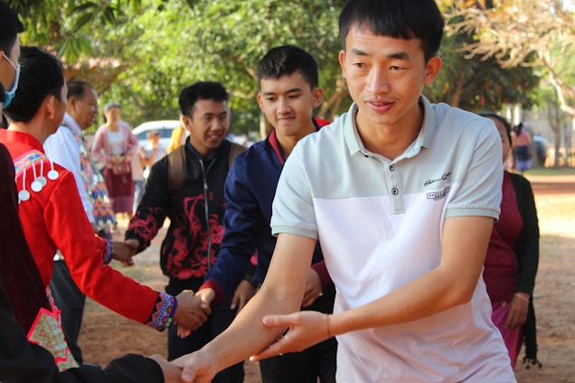 A group of people, including men and women, are gathered outdoors, shaking hands and engaging in conversation. They are dressed in a mix of traditional and casual clothing. Trees and natural elements create a vibrant background, suggesting a community or social event.