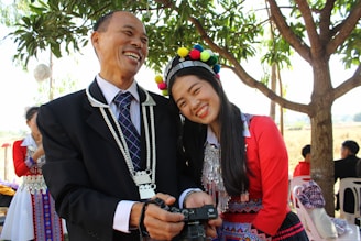 A man and a woman dressed in traditional clothing are joyfully looking at a camera. The man is wearing a black suit with a colorful tie, while the woman is wearing a vibrant outfit with geometric patterns and a headpiece with colorful pom-poms. They are standing outdoors under a tree with bright green leaves.