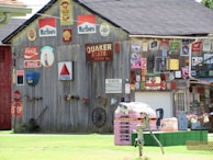 A wooden building covered with vintage signs and advertisements, including brands like Marlboro, Shell, Coca-Cola, and Citgo. There are multiple stop signs, a Quaker State motor oil sign, and various other metal signs and memorabilia. A pile of construction materials sits on the grass, with a water pump nearby and a garage door in the background.