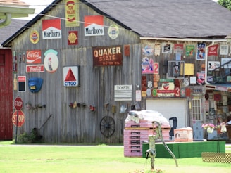 A wooden building covered with vintage signs and advertisements, including brands like Marlboro, Shell, Coca-Cola, and Citgo. There are multiple stop signs, a Quaker State motor oil sign, and various other metal signs and memorabilia. A pile of construction materials sits on the grass, with a water pump nearby and a garage door in the background.