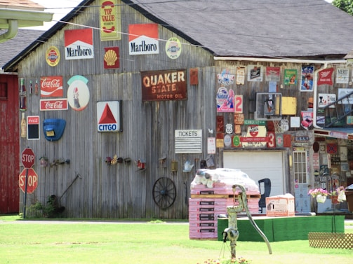 A wooden building covered with vintage signs and advertisements, including brands like Marlboro, Shell, Coca-Cola, and Citgo. There are multiple stop signs, a Quaker State motor oil sign, and various other metal signs and memorabilia. A pile of construction materials sits on the grass, with a water pump nearby and a garage door in the background.