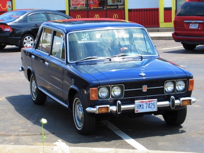 A vintage blue car with a boxy design is parked in a parking lot. The car has Ohio license plates and is situated in front of a fast-food restaurant with yellow and red accents. Other modern cars are parked nearby.