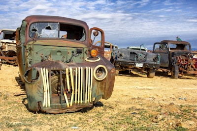 An array of old, rusted truck carcasses is scattered across a desolate, sandy area. The trucks are missing parts and exhibit extensive signs of decay, with broken windshields and weathered bodies. The scene suggests abandonment and highlights the passage of time.