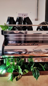 In a cafe setting, a sleek silver espresso machine with the brand name 'strada' is prominently displayed. On top of the machine are several black ceramic coffee pots. Below it, vibrant green leafy plants add a touch of nature against the metallic background.