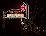 A neon sign reading 'Fireside' is prominently displayed against a dark night background. Below the main sign, a message board reads 'We celebrate unity, compassion, kindness.' The scene is decorated with small twinkling lights illuminating the trees and street beneath the sign. Cars and people are visible along the dimly lit street.