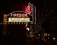 A neon sign reading 'Fireside' is prominently displayed against a dark night background. Below the main sign, a message board reads 'We celebrate unity, compassion, kindness.' The scene is decorated with small twinkling lights illuminating the trees and street beneath the sign. Cars and people are visible along the dimly lit street.