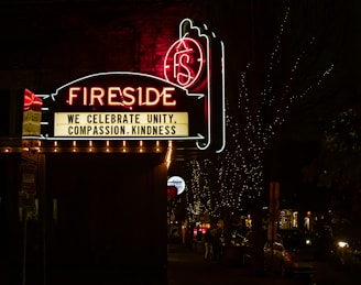 A neon sign reading 'Fireside' is prominently displayed against a dark night background. Below the main sign, a message board reads 'We celebrate unity, compassion, kindness.' The scene is decorated with small twinkling lights illuminating the trees and street beneath the sign. Cars and people are visible along the dimly lit street.