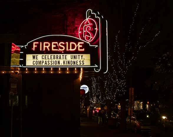 A neon sign reading 'Fireside' is prominently displayed against a dark night background. Below the main sign, a message board reads 'We celebrate unity, compassion, kindness.' The scene is decorated with small twinkling lights illuminating the trees and street beneath the sign. Cars and people are visible along the dimly lit street.