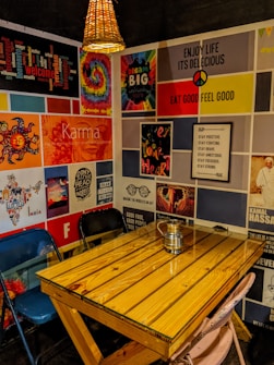 A cozy corner with a wooden table and mismatched chairs, surrounded by a vibrant collage of posters on the walls. The posters feature motivational quotes, colorful artwork, and cultural references. A wicker lampshade hangs above the table, casting a warm glow. A metal jug sits in the center of the table.