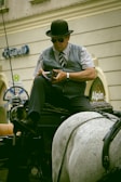 A man wearing a bowler hat and sunglasses sits on a horse-drawn carriage. He is dressed in a vest, tie, and shirt, holding a mobile device in his hand. The background shows part of a building with signs and arched windows.