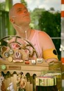 A person wearing a sleeveless top and a nasal cannula is sitting in front of a decorative wooden apparatus adorned with various ornaments, including a crown symbol with the word 'Vienna', miniature figurines, ladybug decorations, and several pins or badges. The apparatus features a 'Danke' sign, suggesting a gesture of gratitude, possibly related to donations.