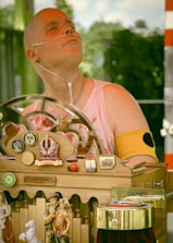 A person wearing a sleeveless top and a nasal cannula is sitting in front of a decorative wooden apparatus adorned with various ornaments, including a crown symbol with the word 'Vienna', miniature figurines, ladybug decorations, and several pins or badges. The apparatus features a 'Danke' sign, suggesting a gesture of gratitude, possibly related to donations.