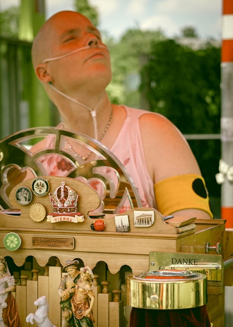 A person wearing a sleeveless top and a nasal cannula is sitting in front of a decorative wooden apparatus adorned with various ornaments, including a crown symbol with the word 'Vienna', miniature figurines, ladybug decorations, and several pins or badges. The apparatus features a 'Danke' sign, suggesting a gesture of gratitude, possibly related to donations.
