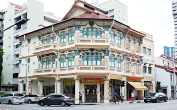 A charming and ornate two-story shophouse with intricate architectural details, featuring colorful floral designs and traditional wooden shutters. The building sits at a busy street corner and includes commercial signs in Chinese characters. Modern cars and a person on a bicycle are visible on the street.