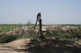 A manual water pump stands in a rural landscape surrounded by green fields and shrubs under a clear blue sky. The ground around the pump is earthy and uneven, with patches of grass and vegetation visible.
