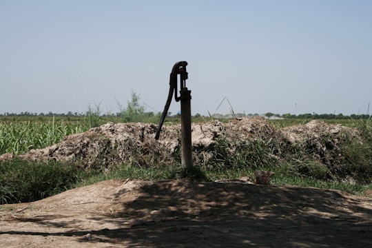 A manual water pump stands in a rural landscape surrounded by green fields and shrubs under a clear blue sky. The ground around the pump is earthy and uneven, with patches of grass and vegetation visible.