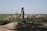 A manual water pump stands in a rural landscape surrounded by green fields and shrubs under a clear blue sky. The ground around the pump is earthy and uneven, with patches of grass and vegetation visible.