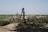 A manual water pump stands in a rural landscape surrounded by green fields and shrubs under a clear blue sky. The ground around the pump is earthy and uneven, with patches of grass and vegetation visible.