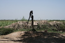 A manual water pump stands in a rural landscape surrounded by green fields and shrubs under a clear blue sky. The ground around the pump is earthy and uneven, with patches of grass and vegetation visible.