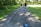 A family of three, consisting of two adults and a child, walks along a paved path surrounded by lush greenery and tall trees. The child is holding hands with both adults. A blue hat is lying on the path in the foreground.