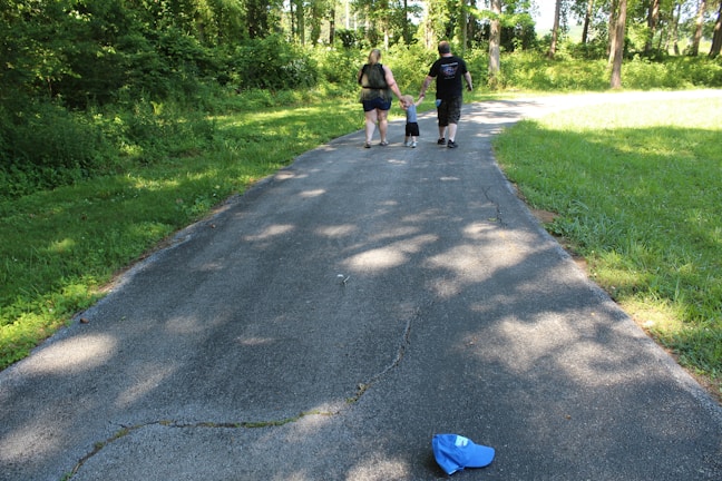 A family of three, consisting of two adults and a child, walks along a paved path surrounded by lush greenery and tall trees. The child is holding hands with both adults. A blue hat is lying on the path in the foreground.
