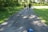 A family of three, consisting of two adults and a child, walks along a paved path surrounded by lush greenery and tall trees. The child is holding hands with both adults. A blue hat is lying on the path in the foreground.