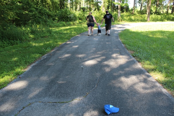 A family of three, consisting of two adults and a child, walks along a paved path surrounded by lush greenery and tall trees. The child is holding hands with both adults. A blue hat is lying on the path in the foreground.
