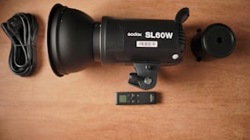 A black Godox SL60W studio light is positioned on a wooden surface. Next to the light, there is a coiled power cable, a remote control, and a cylindrical attachment.