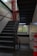A dimly lit stairwell with black metal steps and railings. Red pipes are visible on the left wall, and a window with a grid pattern allows limited natural light into the space. The walls are painted white, and a red and white striped safety bollard is situated at the bottom right corner.