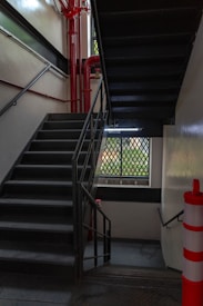 A dimly lit stairwell with black metal steps and railings. Red pipes are visible on the left wall, and a window with a grid pattern allows limited natural light into the space. The walls are painted white, and a red and white striped safety bollard is situated at the bottom right corner.