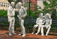 Four white sculptures of human figures are placed in a park setting. Two figures are standing, engaged in conversation, while the other two are sitting on a bench, appearing relaxed and interacting with each other. The background shows green foliage and urban buildings with various signs and flags visible.