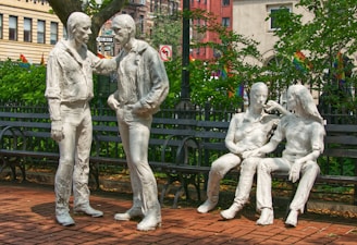 Four white sculptures of human figures are placed in a park setting. Two figures are standing, engaged in conversation, while the other two are sitting on a bench, appearing relaxed and interacting with each other. The background shows green foliage and urban buildings with various signs and flags visible.
