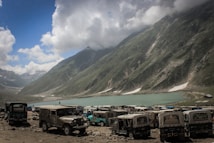 A scenic mountain landscape with a serene lake nestled between lush green hills. Several jeeps are parked on the rocky foreground, while fluffy clouds float in the clear blue sky.