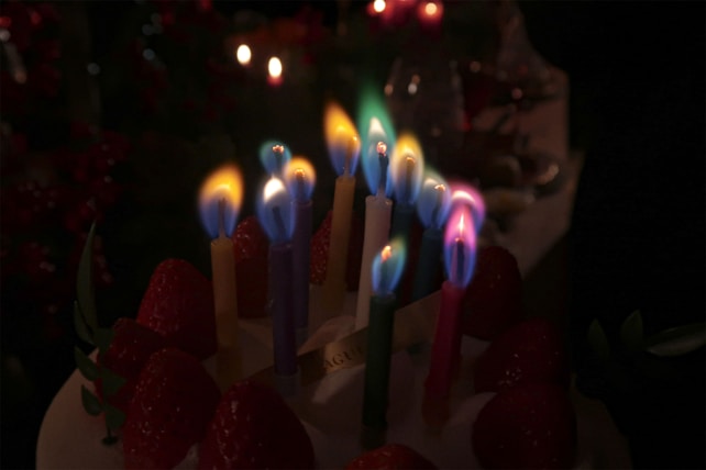 A dark background highlights a cake topped with colorful, lit candles surrounded by strawberries. The candles emit flames in shades of blue, green, and white, creating a festive atmosphere.