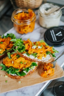 Open-faced sandwiches topped with leafy greens, sliced orange vegetable, and a spread on a piece of parchment paper. A jar filled with more of the orange vegetable and another with a creamy spread are nearby.