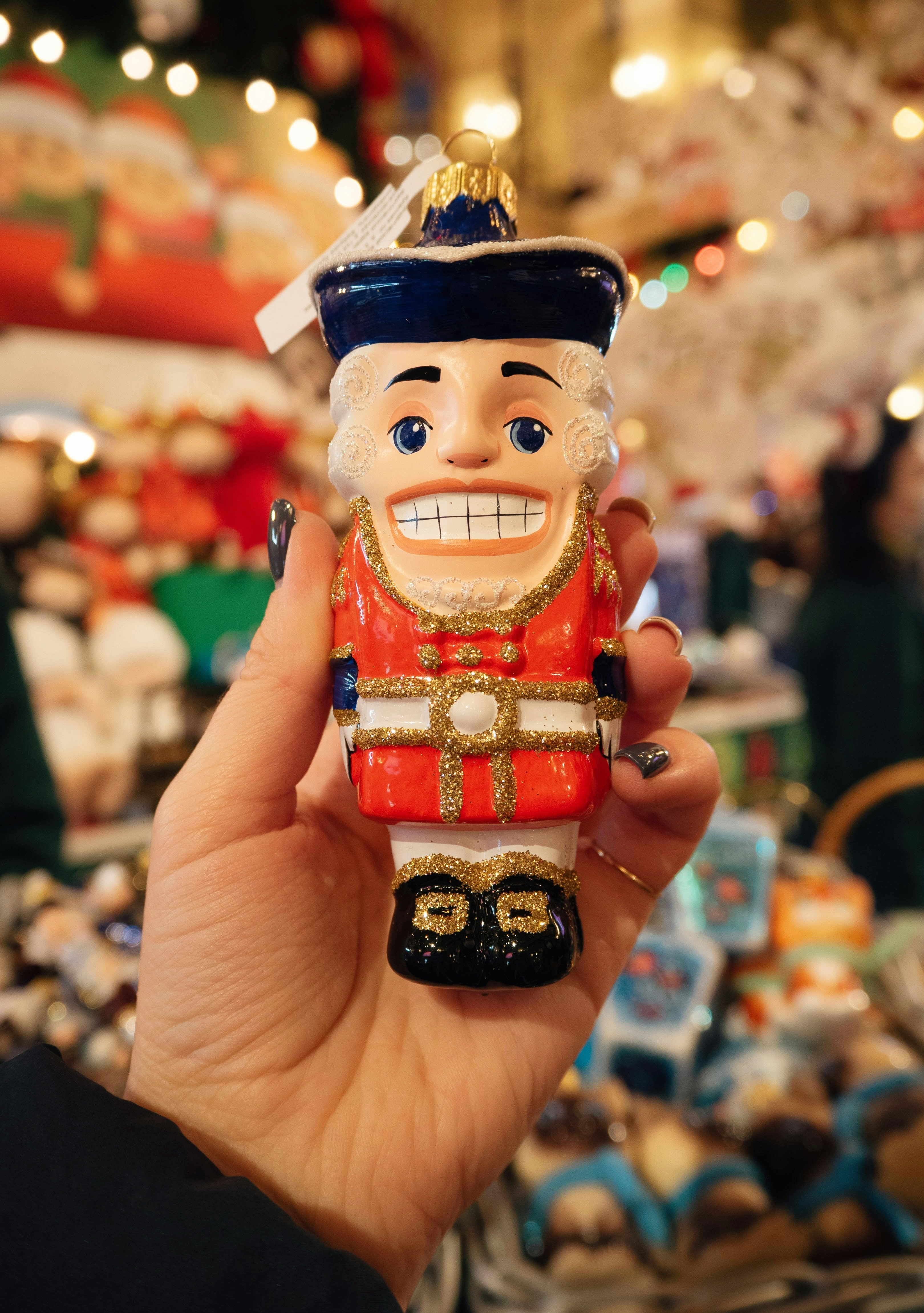 Close-up photograph of a glossy nutcracker ornament held by a hand, with warm Christmas market lights blurred in the background.
