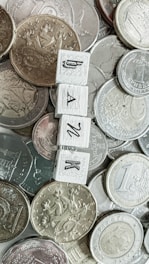 Various coins from different currencies are scattered in a pile. In the center, four wooden tiles with letters spelling 'BANK' are arranged vertically.