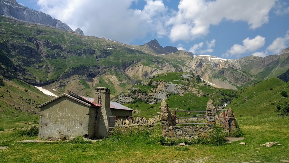 gray concrete house on green grass field near mountain under white clouds during daytime