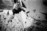 A person is engaged in indoor rock climbing on a bouldering wall. The wall is equipped with various holds and features, and the climber is reaching upward, clad in climbing attire. The scene is captured in black and white, emphasizing the textures and shapes of the climbing holds and the geometric angles of the wall.