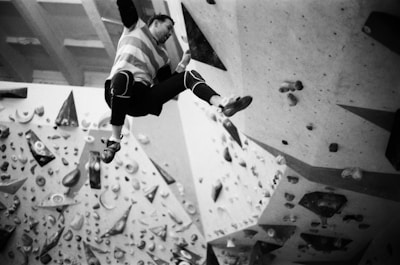 A person is engaged in indoor rock climbing on a bouldering wall. The wall is equipped with various holds and features, and the climber is reaching upward, clad in climbing attire. The scene is captured in black and white, emphasizing the textures and shapes of the climbing holds and the geometric angles of the wall.