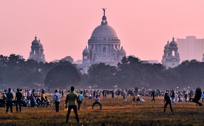 A large open field is bustling with people engaged in various activities, such as playing cricket. In the background, an impressive neoclassical building with a prominent dome and intricate architecture is visible against a dusky pink sky. Surrounding the field are trees, adding to the lively atmosphere of the scene.