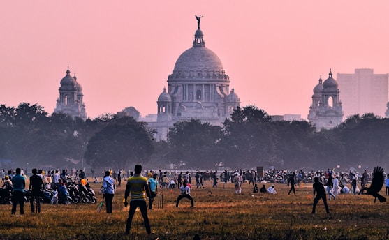 A large open field is bustling with people engaged in various activities, such as playing cricket. In the background, an impressive neoclassical building with a prominent dome and intricate architecture is visible against a dusky pink sky. Surrounding the field are trees, adding to the lively atmosphere of the scene.
