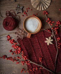A cozy scene featuring a cup of frothy coffee surrounded by autumnal decor. A knitted maroon fabric provides a warm backdrop, accented by a wooden snowflake ornament and red berries on branches. Additional elements include a ball of maroon yarn and rustic star-shaped decorations, creating a warm and inviting atmosphere.