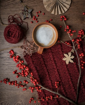 A cozy scene featuring a cup of frothy coffee surrounded by autumnal decor. A knitted maroon fabric provides a warm backdrop, accented by a wooden snowflake ornament and red berries on branches. Additional elements include a ball of maroon yarn and rustic star-shaped decorations, creating a warm and inviting atmosphere.
