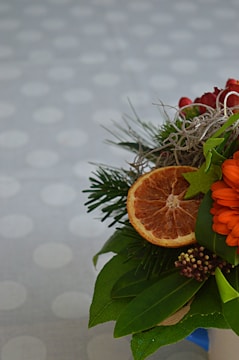 A vibrant floral arrangement featuring an orange flower, dry orange slice, and various green leaves on a dotted, blurred background. The arrangement includes greenery like pine branches and small red berries, alongside curled decorative elements.