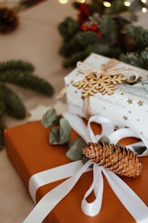 Beautifully wrapped gifts on a table, adorned with natural elements such as pinecones and green foliage. One of the boxes is wrapped in a star-patterned paper with a ribbon and a wooden snowflake decoration. Soft lighting creates a cozy atmosphere, enhanced by the blurred greens and reds in the background.