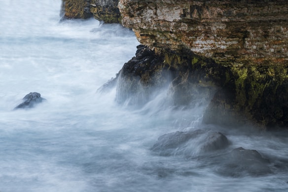 Waves crash against rugged cliffs, creating a misty atmosphere. The rocks are dark and jagged, partially covered in green moss, and the water appears turbulent and frothy.