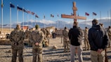 A group of soldiers, dressed in camouflaged uniforms, stand on gravel in a military formation facing a large wooden cross. Several international flags are flying in the background next to a mountainous landscape. The sky is clear with a few clouds.